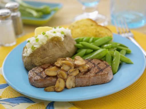 Fleischstück mit Pilzen, Kartoffel, grünem Gemüse und einem Stück Brot auf einem blauen Teller.