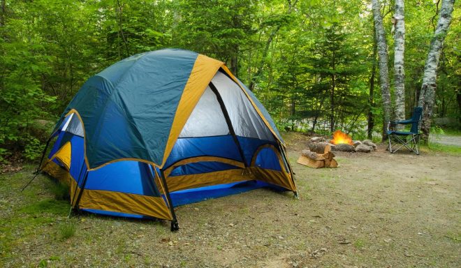 Buntes Zelt in einem Wald, neben einem Lagerfeuer auf einem sandigen Boden.