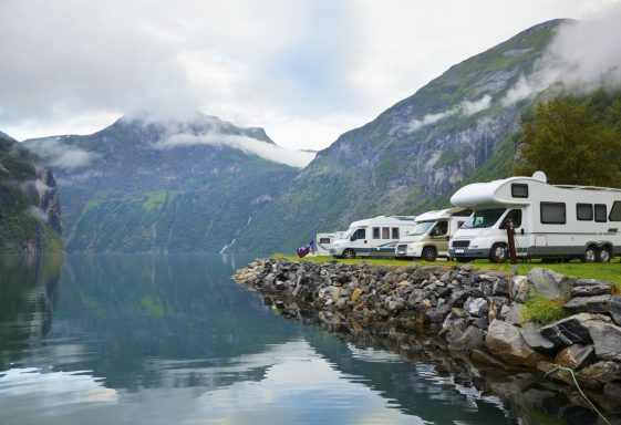 Campingfahrzeuge am ruhigen Wasser eines Fjords, umgeben von Bergen und Wolken.