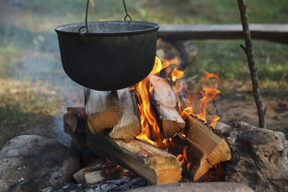 Kessel über einem lodernden Feuer, umgeben von Holzscheiten.