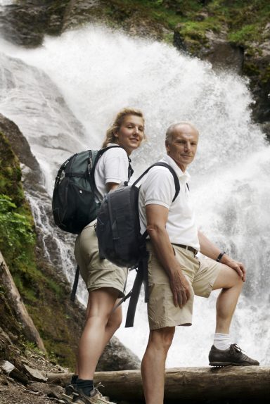 Ein Mann und eine Frau stehen vor einem Wasserfall, beide in Wanderkleidung.