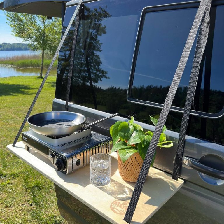 Campingküche mit Pfanne, Gasherd und Pflanze, vor einem schwarzen Wohnmobil am Wasser.