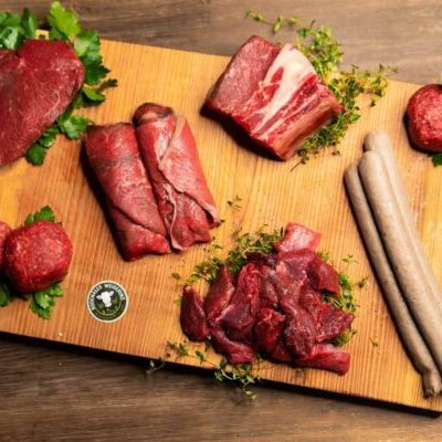 Verschiedene Fleischsorten und Wurst auf einem Holzbrett, umgeben von Kräutern.