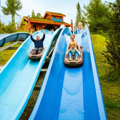 Zwei Rutschen, auf denen Menschen fahren, umgeben von Bäumen und einem Spielplatz im Hintergrund.