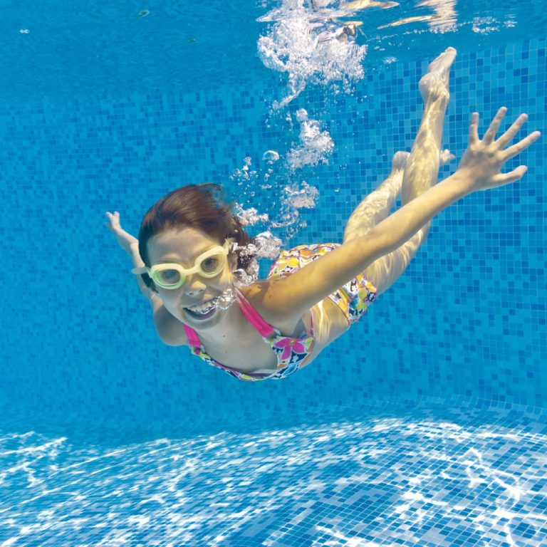 Ein Kind schwimmt fröhlich unter Wasser, trägt eine Schwimmbrille und bunte Badebekleidung.